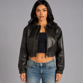 A woman modeling a cropped black faux-leather jacket with a collar and front zipper, paired with a black crop top and blue jeans, against a solid gray studio background.