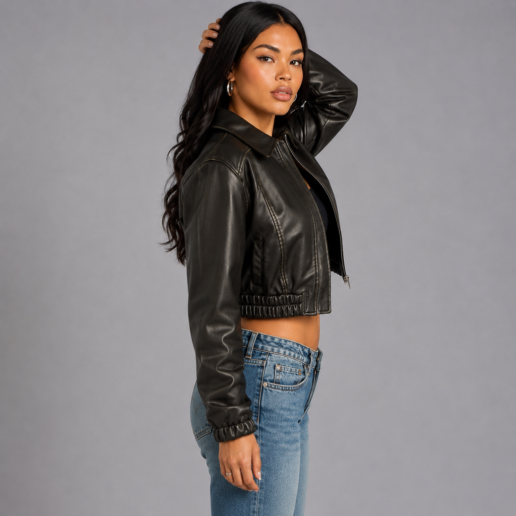 A woman modeling a cropped black faux-leather moto jacket with a collar and front zipper, paired with blue jeans, against a solid gray studio background.