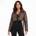 A woman modeling a cropped brown faux-leather moto jacket with asymmetrical zippers and a side tie detail, paired with black pants against a plain white background.