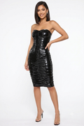 A woman modeling a strapless black ruched midi dress with a glossy, patent-like finish, paired with clear high-heeled sandals, against a plain white studio background.