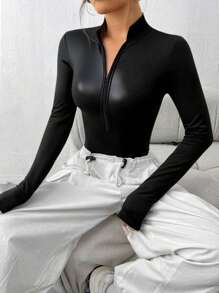 A woman wearing a black long-sleeve mock-neck zip-up top and white pants, seated on a light-colored sofa.